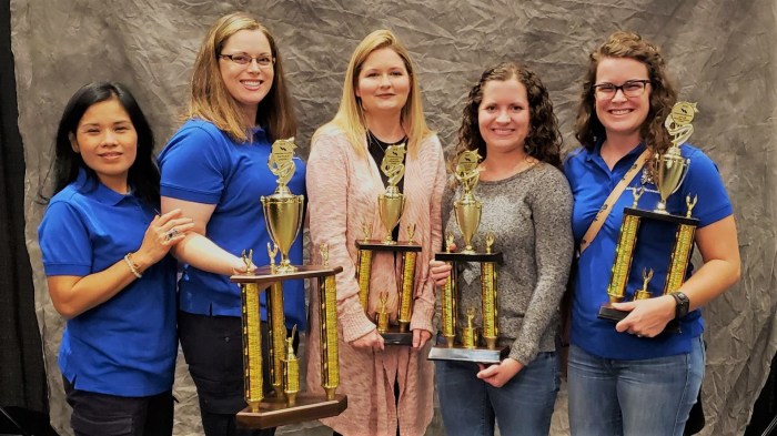 5 utility employees, 3 dressed in blue polo shirts and 2 dressed in casual office clothes hold 4 very large brass-finish and wood trophies