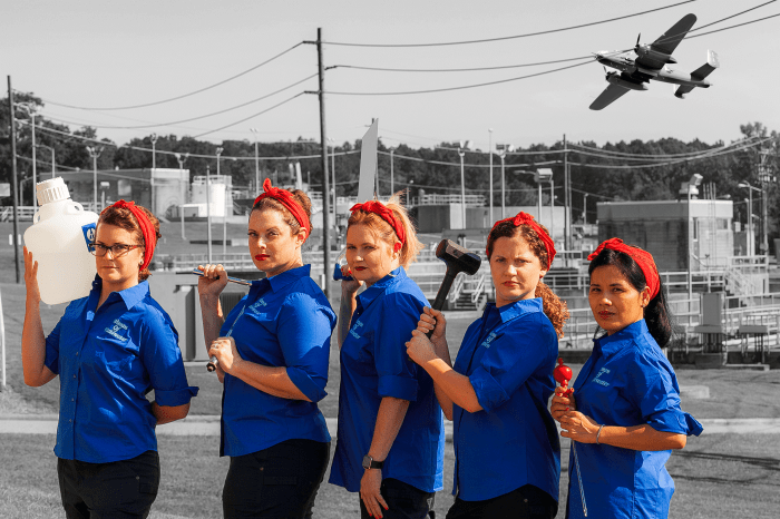 5 utility employees in blue shirts and red bandana headbands stand in a row holding various tools as they pose as Rosie-the-Riveter against a black and white photo of a water treatment plant with a vintage bomber aircraft flies by overhead