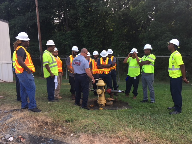over a dozen utility technicians wearing either yellow or orange safety vests and white hard hats stand on grassy area, gather around a yellow fire hydrant; in a blue shirt with a fire department logo on the back, a fire department employee demonstrates use of the hydrant with a large pipe wrench