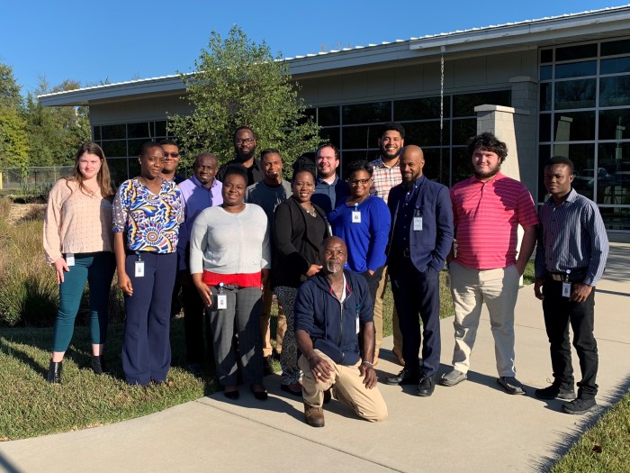 15 utility employees in casual office attire pose for a group photo outside a one-story building with a wall of windows