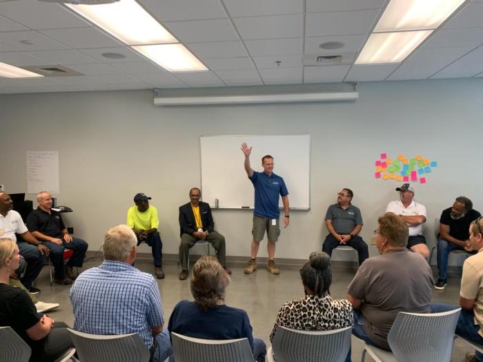 over a dozen utility employees and technicians gather in a grey conference room seated in a circle of chairs; 1 technician in blue polo shirt and brown shorts stands by a large whiteboard as he waves to the group