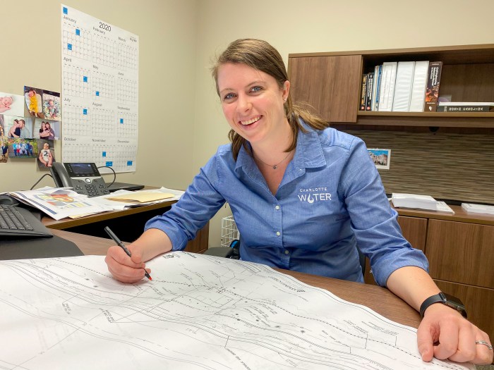 utility engineer sits at desk in office wearing blue blouse with Charlotte Water logo on her shirt as she reviews plans on a large white schematic unrolled, desktop