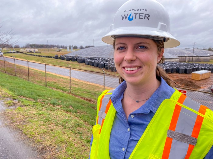 utility engineer poses for photo wearing white hard hat, yellow safety vest, blue blouse and necklace stands outside in front of several concrete domed water treatment ponds and over a hundred black pallet sized, cube-shaped storage bags