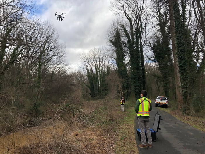 standing along a wood-lined roadway next to a creek, 2 utility technicians wearing yellow safety vests stand near a white pick up truck and large black trunk as they look up a drone in the sky