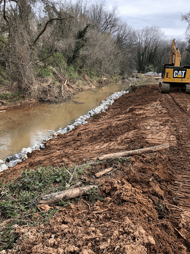 dirt roadway and embankment run alongside a wood-lined creek, in between is a border of gravel and rolling away in the distance is a yellow CAT excavator