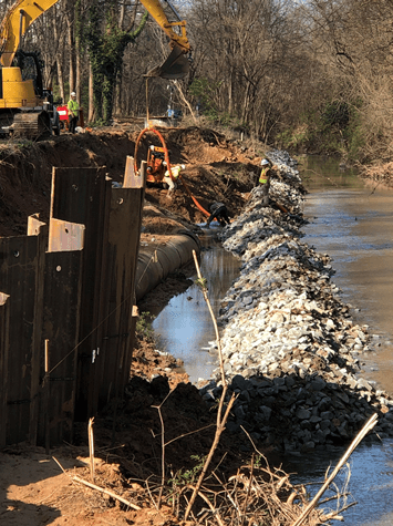corrugated steel retention wall is installed along a long dirt trench, next to it is a long barrier of gravel running alongside a stream, in the distance is a yellow excavator digging dirt out of the trench and 4 utilit workers in yellow safety vests and hard hats manning an orange hose and pumping water out of the trench