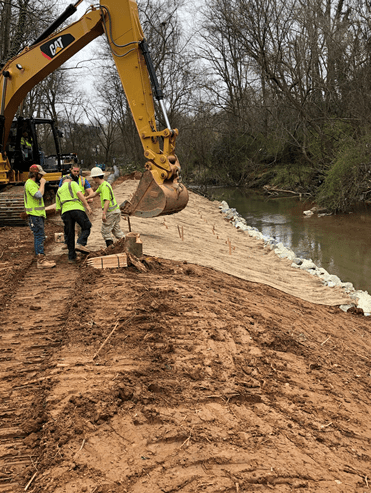 4 utility technicians wearing yellow safety shirts stand near a yellow excavator arm along the dirt road and embankment built alongside a wood-lined creek