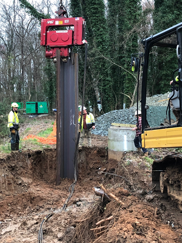 yellow excavator outfitted with a red, rectangular jackhammer style tool pounds a section of brown corrugated steel panel into the side of a dirt trench, 2 utility technicians wearing yellow safety jackets and white hard hats stand nearby, inspecting