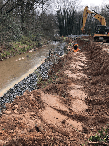 yellow excavator in the distance has dug a long trench in the dirt, a line of gravel follows alongside