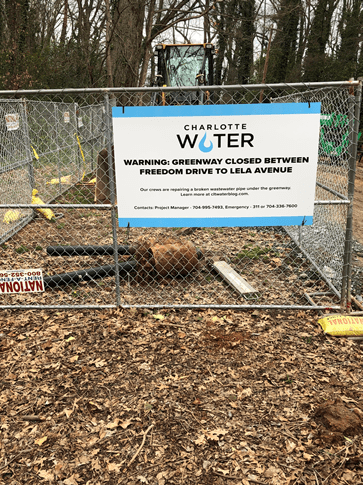 in a wooded area, chain link fence displays a large white sign with blue trim and Charlotte Water logo, reads in black font Warning Greenway Closed Between Freedom Drive to Leila Avenue