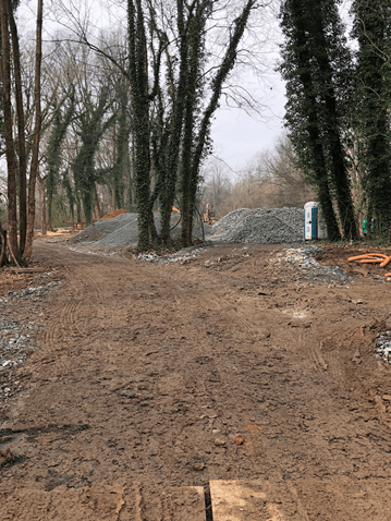 dirt roadway runs past sections of trees and large piles of gravel, blue and white porta-potty, and a small pile of orange hoses