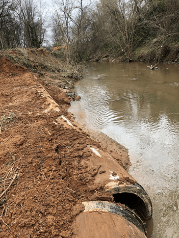 recently cut dirt trench alongside a creek reveals partially buried, old brown steel pipeline with a section that has broken open at a joint