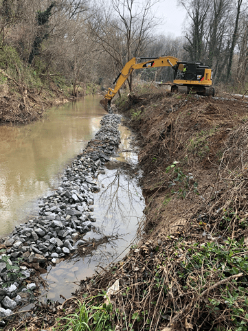 dirt roadway and trench run alongside a wood-lined creek, in between is a long border of gravel; in the distance the arm of a yellow excavator reaches down to adjust the gravel with its bucket