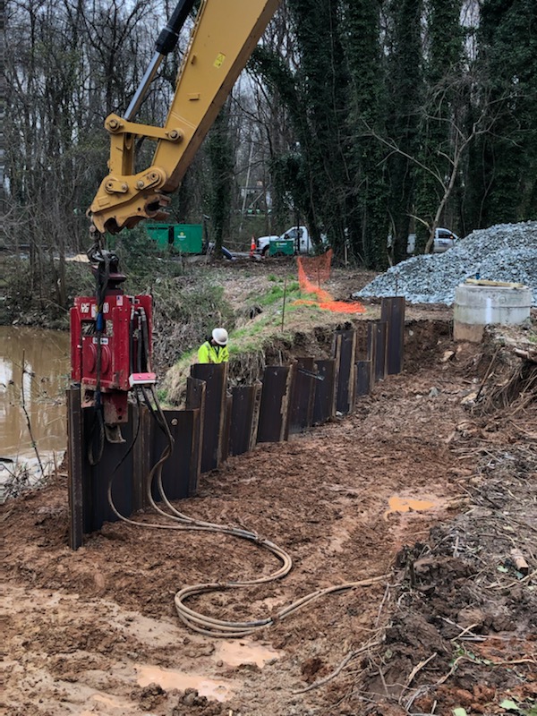 yellow arm of an excavator rigged with a red jack hammer device pounds sections of brown corrugated steel panels into the dirt of a muddy trench, nearby a utility technician in yellow safety shirt and white hard helmet inspects panels previously set in