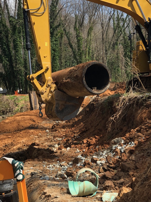 giant yellow excavator balance a section of old steel pipe on its bucket as it lifts it up out of dirt trench