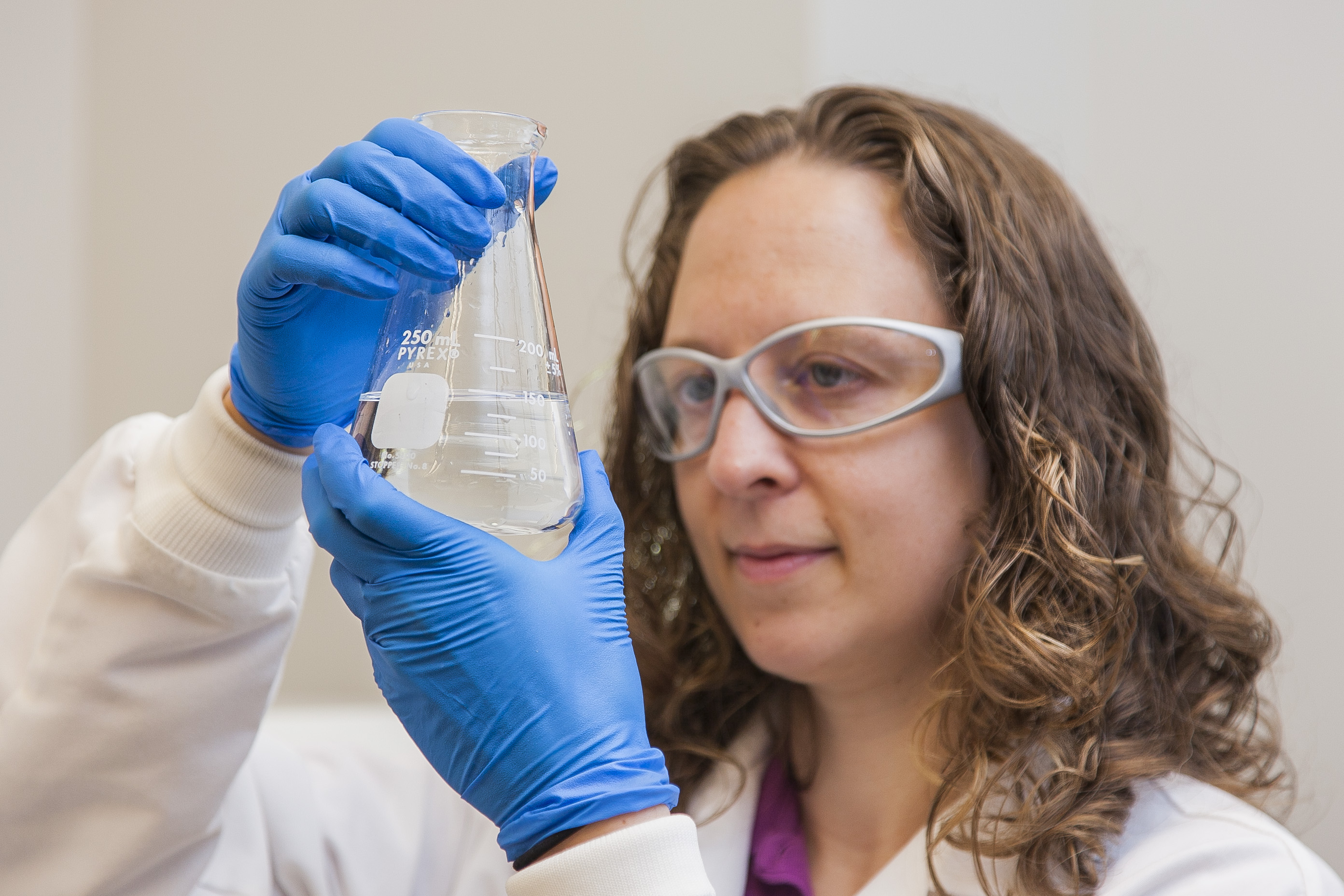 lab technician with long brown wavy hair, grey safety classes, and blue gloves holds up a beaker with clear liquid