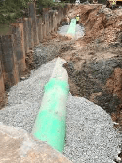 partially covered in gravel, a green pvc pipeline runs through a long muddy trench with a rusty brown corrugated steel retention wall on one side