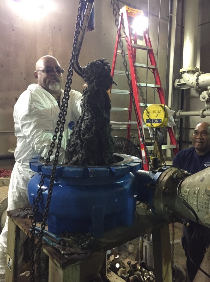 inside a concrete building, a utility technician in white tyvek jumpsuit helps guide a giant hook pull a massive glob of grease and hair out of a blue steel gasket attached to brass and grey valve