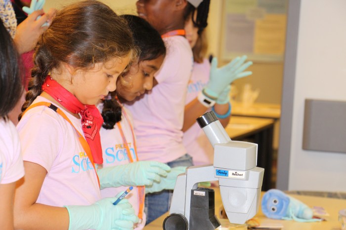 2 school children in pink t-shirts and light blue gloves explore using a grey micro-scope during an education science event