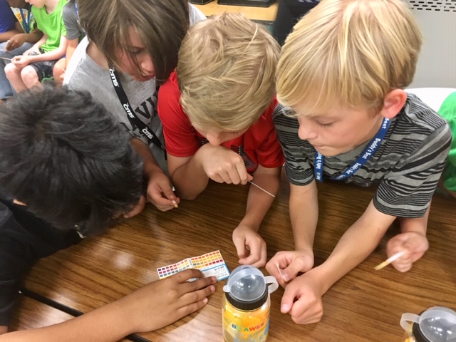 huddled over wooden table, 3 school children holding ph test strips look at the ph test trip key card with their instructor