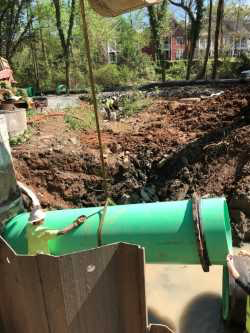 section of large green pvc pipe is lowered into a dirt trench via steel cable as a utility technician in a yellow safety vest and white hard hat helps guide the pipe into place