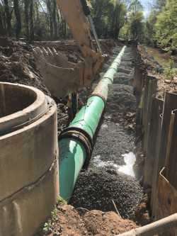 many sections of green pvc pipes connected by brown gaskets rests along a very long dirt trench cut through the ground, lined with metal retention walls, in the foreground is the bucket of a yellow excavator and a round concrete manhole access stack
