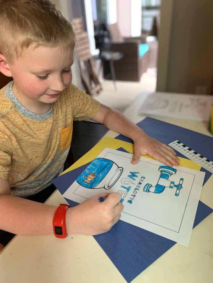 seated at a tan table, a school child in peach t-shirt with grey collar and red wrist watch uses a blue crayon to fill in a coloring book sheet with a water theme