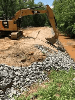 earthen retention wall being shaped by yellow excavator, below it is a line of rocks and a muddy pond