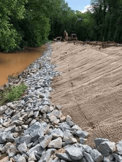 next to a muddy pond, a course of crumbled grey rock runs along the side of an earthen retention retention wall