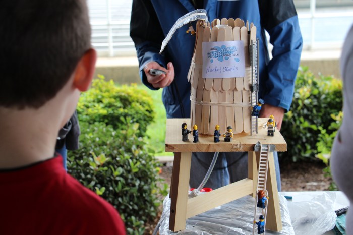 model of a water tower made with wooden tongue depressors and wood decorated with toy plastic ladders, little plastic people is displayed on a table at an outdoor educational exhibition