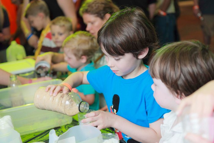 young boy with blue t-shirt pours a peach colored powder from a clear plastic bottle into a cup as other children surround him