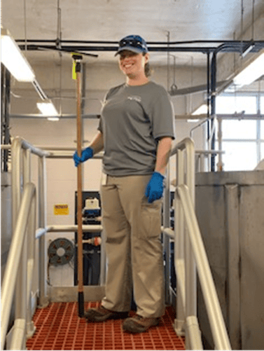 inside a water treatment facility building, a utility technician wearing grey t-shirt, blue baseball cap and blue gloves holds a wooden pole as she stands on brick colored, grated rubber mat on a small platform with metal railings.