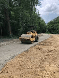 yellow steamroller rolls down a grey grave, tree-lined rural street