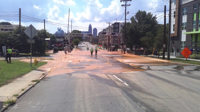 muddy water floods a city street as 2 utility workers in yellow safety vests inspect it by walking through it