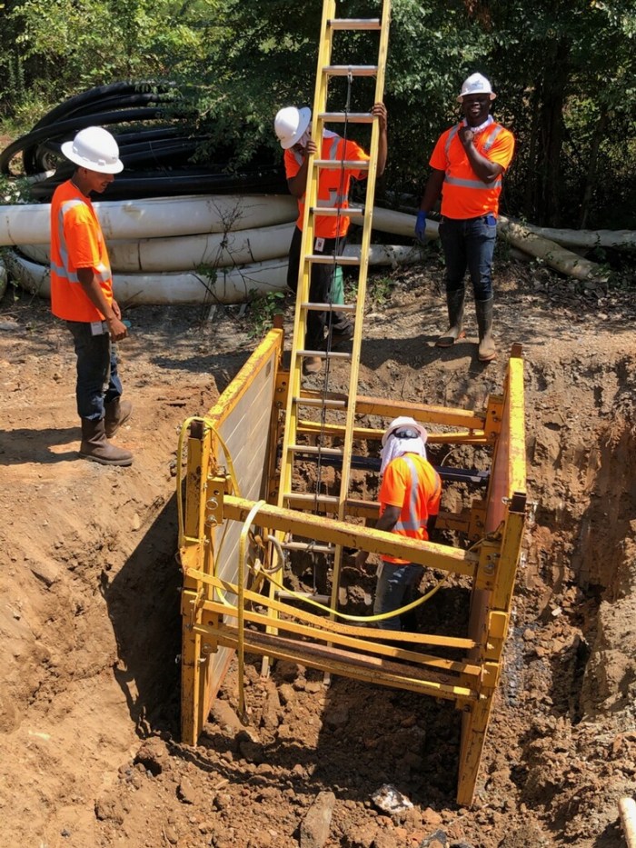 1 utility technician inspects the ground inside a yellow steel frame along with yellow ladder at the bottom of a square dirt pit, above 3 other utility technicians stand next to a pile of white flexible pipes, all wear orange safety t-shirts and white hard hats