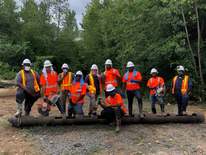 10 utility technicians wearing orange safety shirts and white hard hats, line up in an open space next to a wooded area to pose for a group photo as they stand on section of pipe with cuts in it