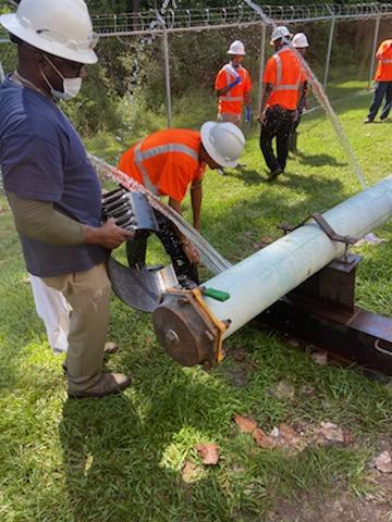 2 utility technicians wearing white hard hats work to position a section of capped white pipe on a stand, behind them on grassy section of chain link fenced lawn are 4 other utility technicians