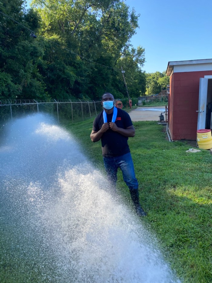 utility employee wearing a black t-shirt, mask and blue towel around his neck stands outside on a grassy lawn next to a spray of water and a small brick sub-station building