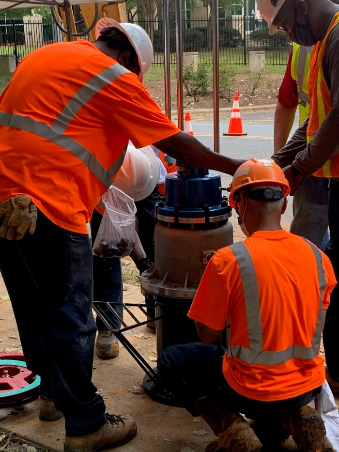 along side a city street with 2 orange traffic cones,5 utility technicians wearing orange safety vests or shirts and hard hats circle around a tan pipe valve stack with a blue cap