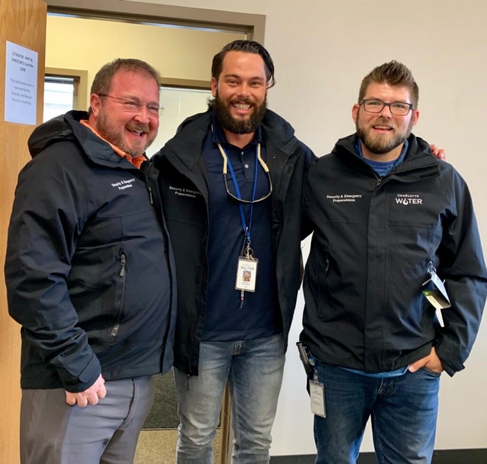 3 utility engineers in dark blue jackets with Charlotte Water logos pose for a group photo in white painted hallway
