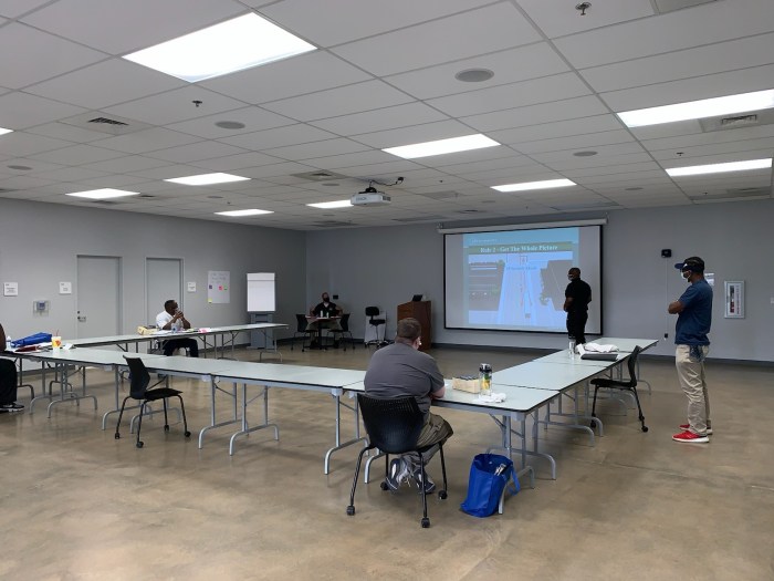 seated and standing around a horseshoe layout of tables and chairs, 5 utility employees watch a ppt slide presentation projected onto a screen in a large, grey and tan conference room