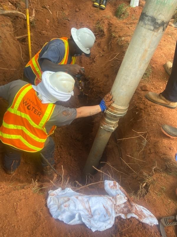 2 utility workers wearing orange safety vests and white hardhats stand in a dirt trench assisting a drill going into the bottom of the trench