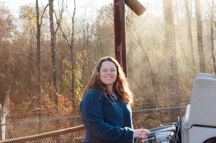against a wooded backdrop, 1 utility engineer stands next to a tall brown pipe shaped vent stack typing on a grey plastic console next to her