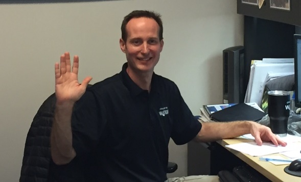 utility employee wearing black polo shirt with Charlotte Water logo waves at the camera while seated at tan and black desk in an office