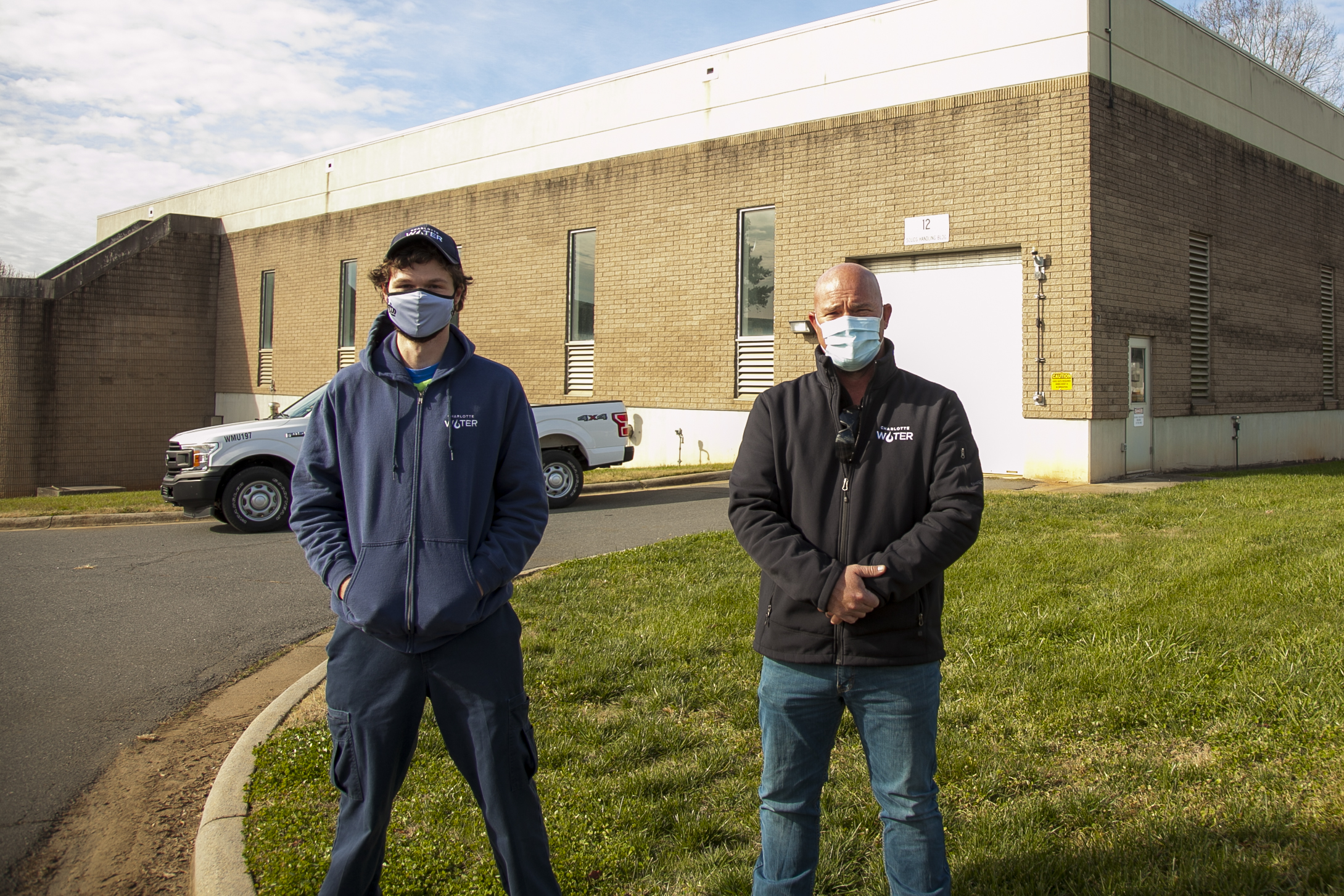 2 utility employees wearing jackets with Charlotte Water logos and masks stand along a grassy curb for a group photon in front of a tan brick with concrete trim two-story building behind them