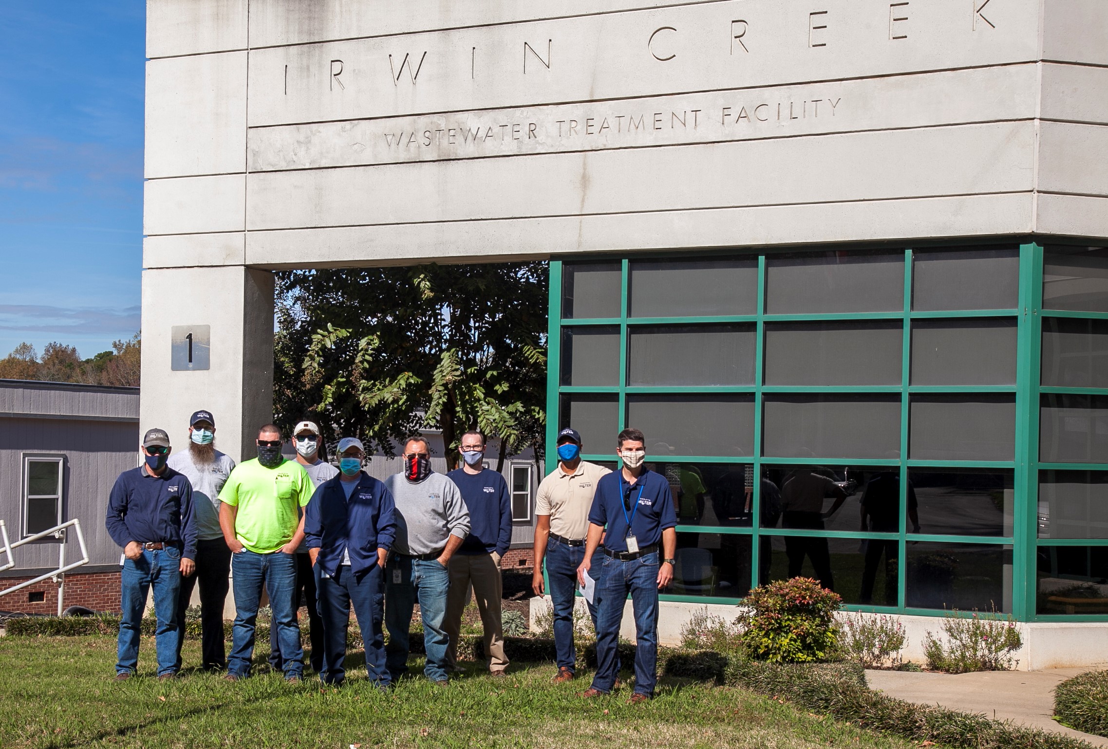 9 utility employee wearing polo shirts, t-shirts and masks stand outside posing for a group photo in front of a concrete building with green framed windows with the name Irwin Creek Wastewater Treatment Facility on it