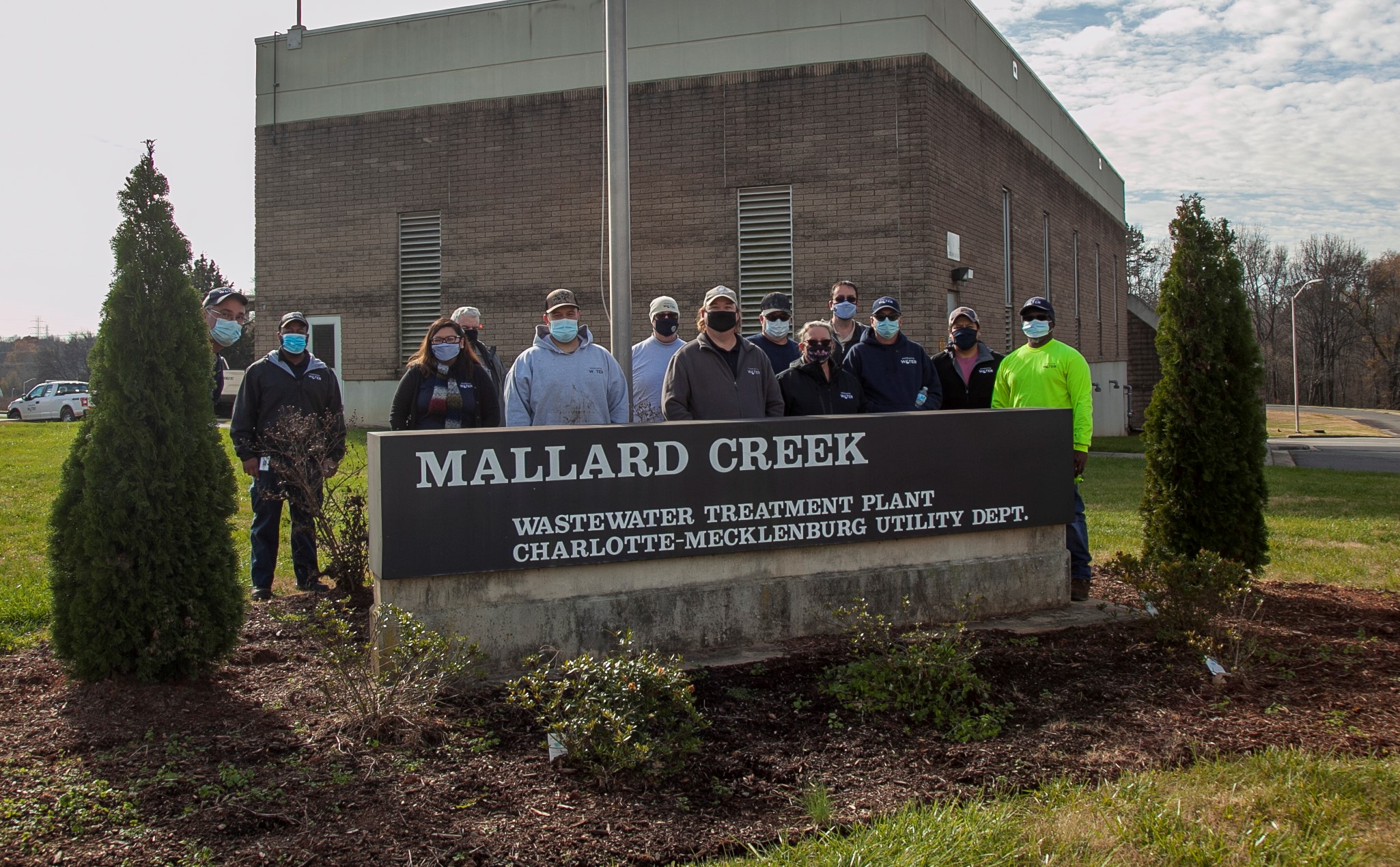 a dozen office and utility employees stand outside wearing office and construction clothes and masks as they pose for a group photo behind a new sign Mallard Creek Wastewater Treatment Plan Charlotte-Mecklenburg Utility Dept, a large brown brick and tan concrete two-story building stands behind them