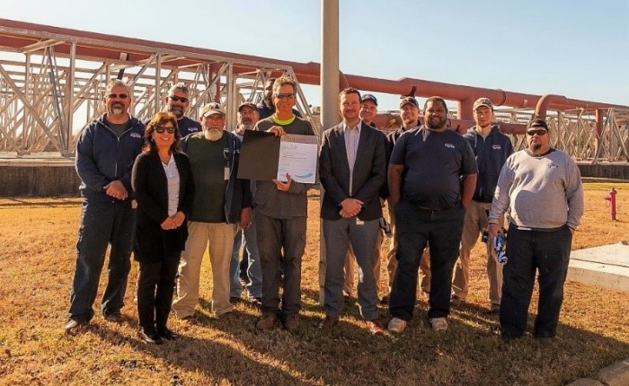 a dozen utility employees dressed in either office attire or utility work clothing stand outside on a grassy area in front of a large scaffolded-style structure supporting 2 large water pipe arrays above; tall man wearing green shirt in the middle of the group holds a brown and white awards plaque