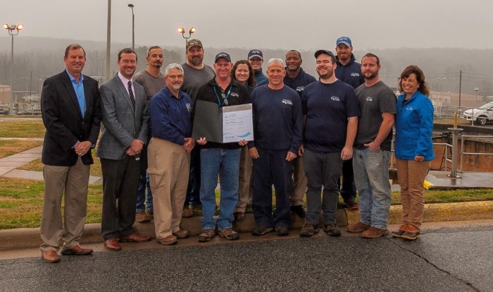 14 utility employees dressed in either office attire or utility work t-shirts stand outside in front of water treatment plant facility, one man in center holds a large white and brown awards plaque