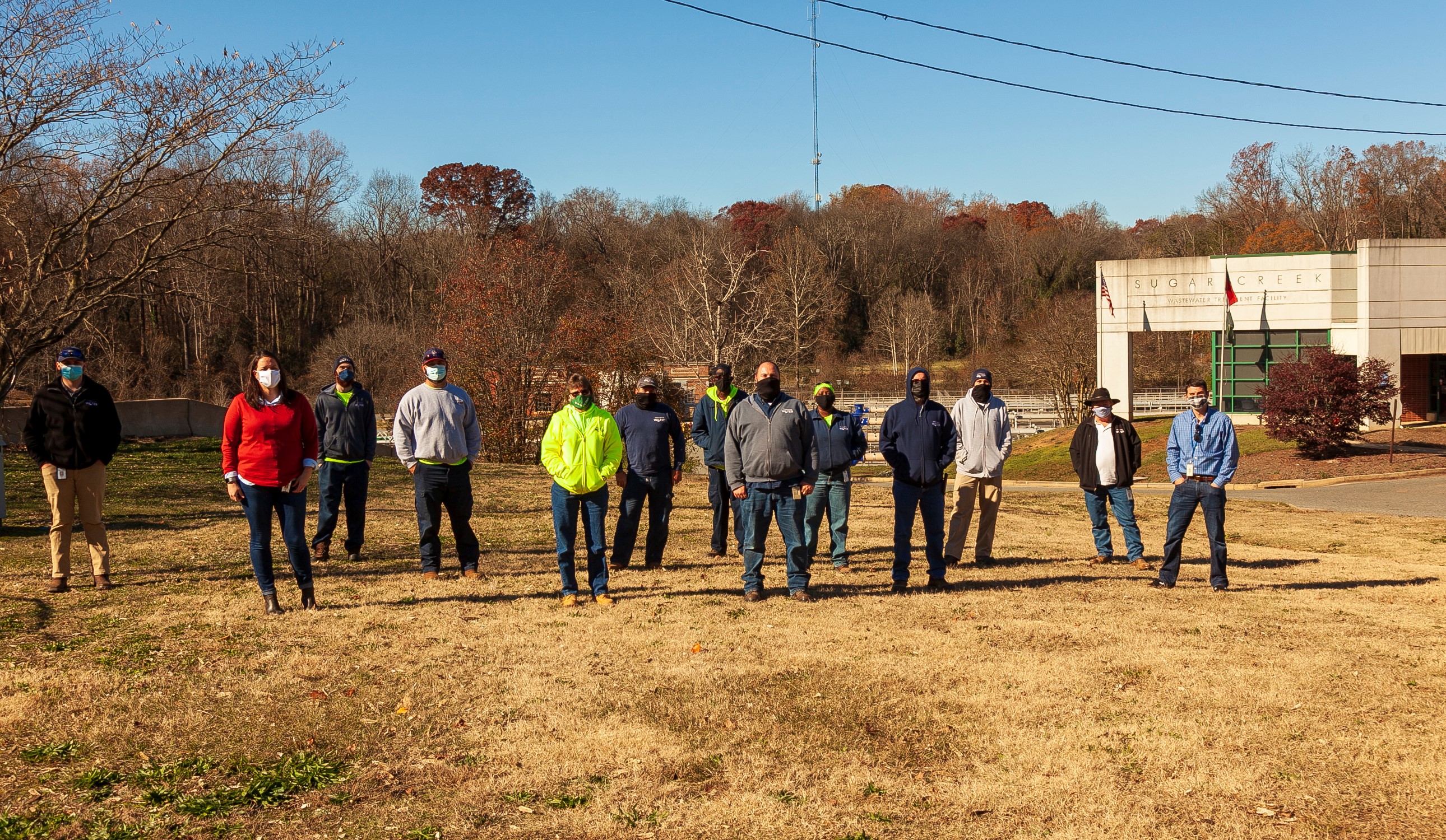 a dozen utility employees and technicians wearing office and construction clothing stand masked outside on brown lawn in front of a late autumn tree-lined creek and a tan two-story building with the name Sugarcreek on its front